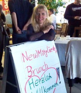 Eine blonde Frau, Anfang 40, lehnt freudig über einem Aufsteller mit der Aufschrift: Neumitglieder-Brunch Herzlich Willkommen!