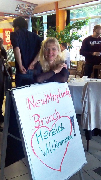 Eine blonde Frau, Anfang 40, lehnt freudig über einem Aufsteller mit der Aufschrift: Neumitglieder-Brunch Herzlich Willkommen!