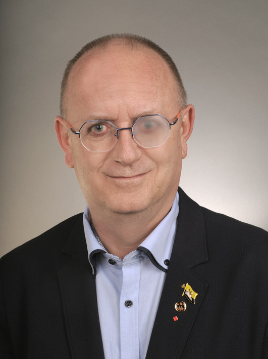 Jörg hat schütteres braunes Haar, trägt eine Brille, linkes Auge mit Milchglas, trägt eine NZUGJcke und blauem Hemd